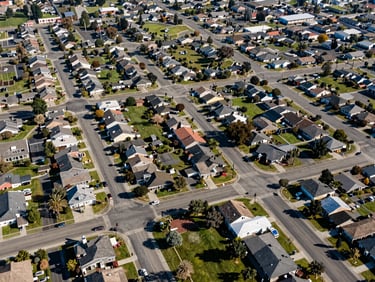 Overhead wide shot of a NorCal suburb at midday — grid of streets, parks, and rooftops showing scale of community, clear daylight, shot from moderate altitude