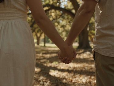 Intimate close-up of a couple holding hands while walking through a sun-drenched oak forest, golden light filtering through trees, deep natural shadows.