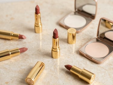 Elegant flat-lay of luxury gold-cased lipsticks and fine powder compacts on a warm beige marble counter, soft daylight