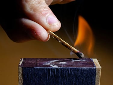 A person striking a wooden match against a matchbox, creating a bright orange flame and smoke.