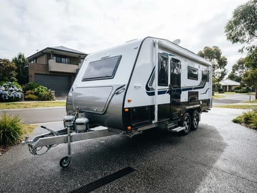 Low-angle heroic wide shot of a pristine caravan parked in a clean Melbourne driveway, water droplets glistening on its glossy surface under bright overcast daylight, 35mm.