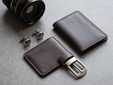 Flat-lay product photography of premium accessories—structured cufflinks, a leather card holder, and a polished metal clasp arranged on a neutral cool-grey surface, diffused daylight from above