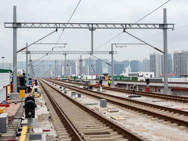 Wide panoramic view of a completed Hong Kong rail corridor and overhead electrification gantries under overcast daylight, finished track bed and signalling equipment visible, environmental scale showing project magnitude