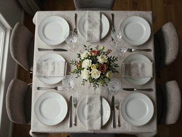 Overhead flat-lay of an intimate private dinner table set for eight — bone china, crystal glasses, folded linen napkins, a single low floral arrangement, neutral window light from the left edge