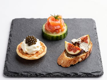 Overhead macro close-up of three delicate appetizers on a slate tile — a blini with crème fraîche and caviar, a cucumber round topped with smoked salmon mousse, a fig crostini with gorgonzola — studio strobe light from directly above, white negative space at edges