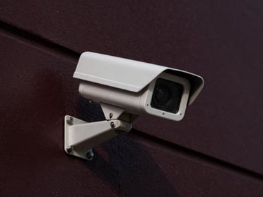 Close-up of a security camera lens on a dark industrial wall, sharp focus, high contrast, dramatic side lighting, deep charcoal and burgundy tones.