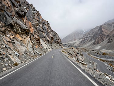 Road-level shot looking ahead on a steep switchback pass in the Karakoram, raw tarmac surface crumbling at the edge, towering rock face to the left, overcast high-altitude light, wide environmental frame showing the road curving upward