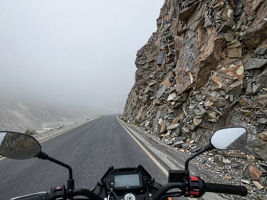 Road-level view from a motorcycle handlebar mount approaching a narrow high-altitude pass in the Karakoram, loose gravel visible on tarmac edges, overcast flat light, sheer rock face filling the right third, road vanishing into grey mist ahead