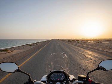 Wide environmental frame from a motorcycle seat looking down the Makran Coastal Highway, flat grey Arabian Sea to the left, cracked tarmac stretching to the horizon, golden-hour light low and warm on the road surface, no vehicles ahead