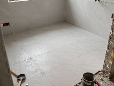 Wide shot of a bathroom mid-tile installation, floor and wall tile partially set, grout lines visible, natural daylight, crew tools on the floor edge of frame, documentary construction photography
