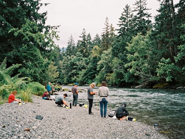 A group of people working along a lush, gravelly riverbank under overcast skies, soft natural daylight, 35mm film style.
