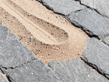 Close-up of quartz sand being swept into paving stone joints, bright natural sunlight, clean texture, crisp outdoor photography, rich slate gray tones.