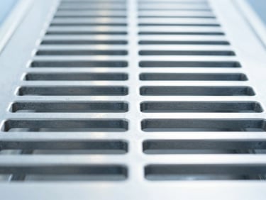 Straight-on macro shot of a pristine, clinical-clean metal ventilation grate with bright daylight reflecting off the polished steel, sharp focus, 35mm lens