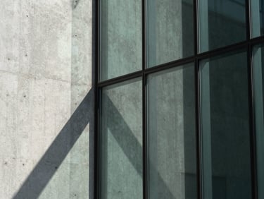 Macro detail of high-performance architectural glazing with sharp daylight shadows, concrete and glass textures, desaturated green accents.