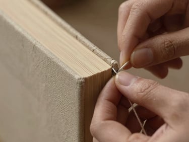 Macro close-up of hands carefully stitching the spine of a linen-bound book with thick thread, warm side lighting highlighting paper fibers