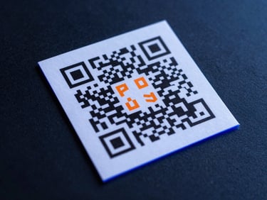 Overhead studio shot of a printed area-announcement flyer with a bold QR code centered, placed on a dark navy surface, cool royal-blue rim light along the edges, warm orange accent highlight on the QR code detail, fine paper texture visible