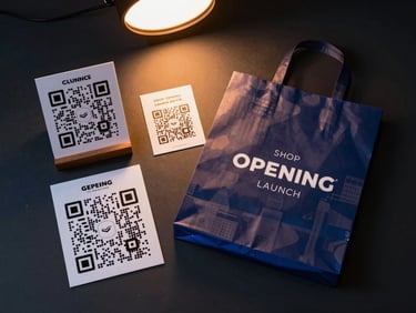 Studio flat-lay of a shop-opening launch kit: standee card, QR sticker sheet, and branded shopping bag arranged on a deep charcoal surface, controlled fill light from above, warm orange glow on the QR code elements, cool blue rim light defining product edges