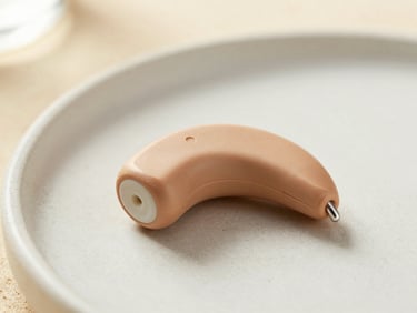 Macro photography of an elegant, ultra-discreet modern hearing aid resting on a soft sand-textured ceramic surface, warm natural lighting, shallow depth of field.