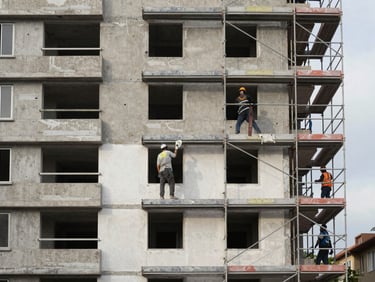 Environmental wide shot of a building facade under partial renovation, scaffolding on the right, workers applying render at mid-height, morning overcast light, raw concrete and finished plaster visible side by side