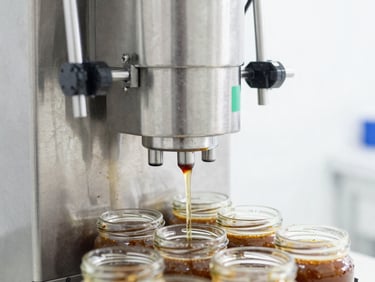 Macro shot of a stainless steel automated liquid filling machine depositing clean sauce into glass jars, high-key lighting, sterile commercial environment