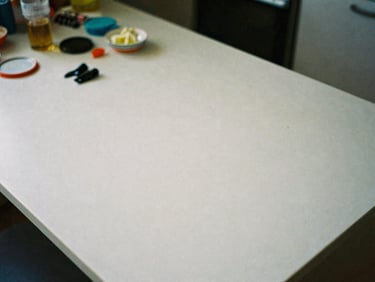 Overhead shot of a messy kitchen table with scattered objects, harsh overhead light, visible film grain, 35mm perspective, raw documentary style.