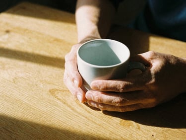 Close-up of hands holding a warm ceramic mug, harsh morning light casting long shadows on a textured wooden table, visible film grain, 35mm perspective.