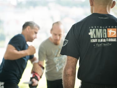 An instructor in a 'Krav Maga Global' shirt observes students practicing self-defense techniques outdoors with a knife.