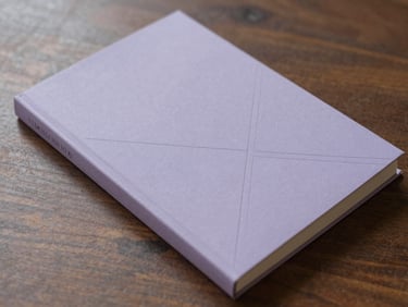 A minimalist book cover with blind-debossed geometric lines on lavender-grey paper, lying flat on a dark wooden table in soft morning light.