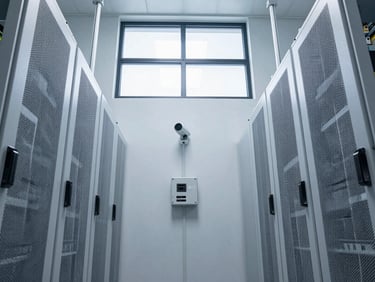 Wide overhead shot of a clinical server room with white-painted walls, mounted security hardware and cable trays, cool north-facing daylight from a high window, no personnel visible