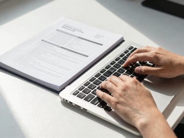Close-up overhead shot of hands typing on a laptop keyboard in a bright office environment, a printed training workbook open beside the laptop, natural daylight from a nearby window casting clean even light across the desk surface, no faces in frame