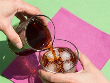 Close-up of hands pouring dark cold brew coffee into a glass filled with ice cubes, bright morning sunlight, vibrant magenta napkin in background.