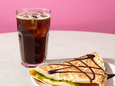 Condensation-covered glass of cold brew coffee next to a warm folded crepe with chocolate drizzle on a light surface, bright daylight, vibrant pink accents