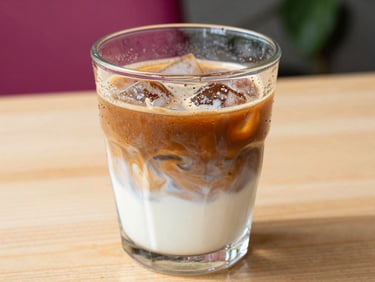 Tight macro shot of a condensation-covered glass of cold brew coffee with swirling milk, sitting on a bright wooden table in warm afternoon daylight, shallow depth of field.