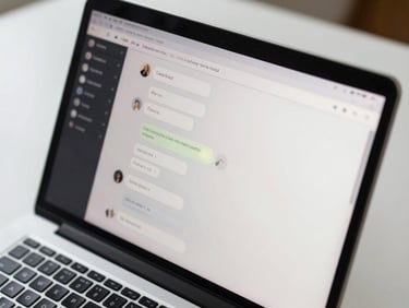 Close-up overhead view of a laptop screen displaying a clean chat interface with message bubbles, glowing softly on a tidy white desk under bright daylight, keyboard partially visible at bottom edge