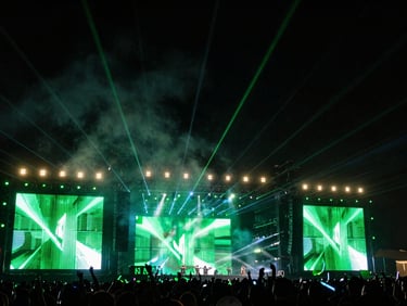 Wide dynamic shot of a massive festival stage with giant LED screens displaying abstract green graphics, heavy smoke, lasers shooting into the night sky, Santiago