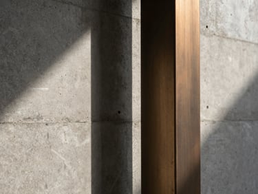 Abstract close-up of a raw concrete wall meeting a brushed bronze structural column, soft natural light casting long linear shadows.