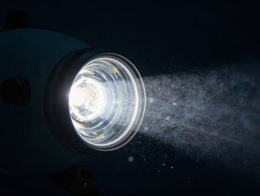 Cinematic close-up of a deep-sea submersible headlight cutting through pitch-black ocean water, biological particles floating in the beam.