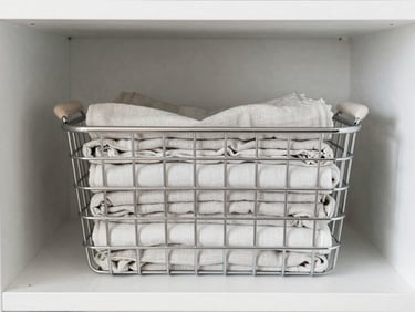 A neat wire basket holding folded linen in a bright, modern closet, soft daylight, desaturated cool tones.