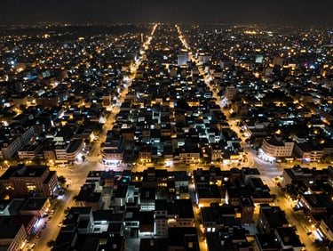 Overhead night aerial view of a city grid with dense street-light patterns, Jaipur rooftop visible in the foreground, wide panoramic composition showing urban scale and connectivity