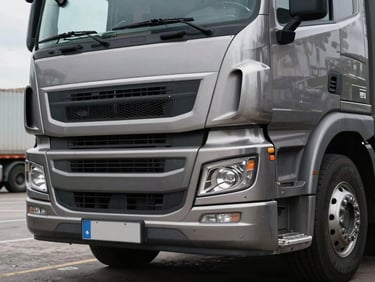 Close-up of a commercial truck front grille and headlights parked at a logistics yard, overcast daylight, sharp metallic detail visible on the bumper