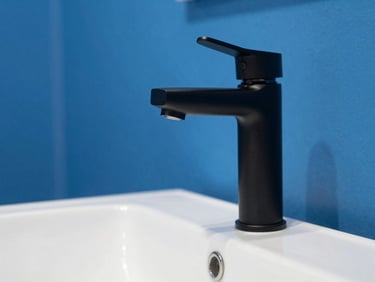 Close-up of a modern matte black mixer tap over a white ceramic basin in a minimalist bathroom, bright cool daylight, bold blue and white tones, crisp clean focus, 35mm lens