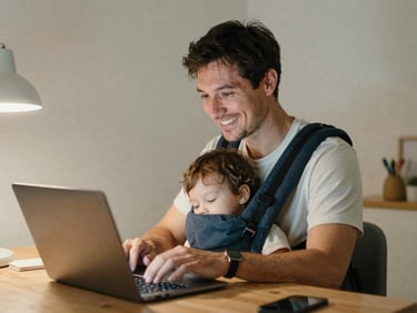 A smiling single father coding on a laptop at night with a sleeping toddler in a carrier, soft ambient desk lamp lighting