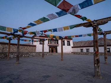 An ancient stone monastery courtyard in Ladakh at dusk, soft blue-hour light, prayer flags gently fluttering, weathered wooden pillars, serene negative space