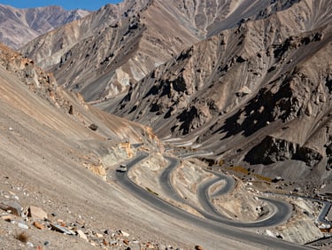 Wide panoramic view of a winding mountain pass in Ladakh, a single small expedition vehicle navigating the massive scale of the bare rocky terrain under harsh high-altitude sunlight.