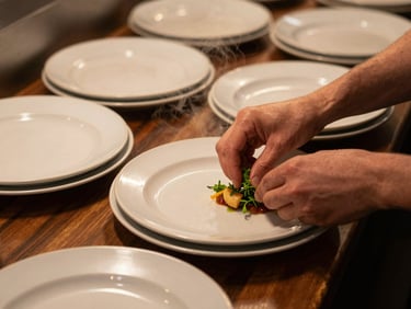 Close overhead shot of a worn restaurant pass during mid-service, plates lined up, hands adjusting garnish, warm tungsten light from above, natural steam rising, dark wood surface beneath