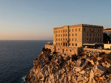 Wide environmental shot of a historic stone hotel perched on a rugged cliff overlooking the deep blue Mediterranean Sea at golden hour, warm light.