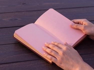 Manos sosteniendo un diario antiguo de cuero, con sutiles detalles en pan de oro en las páginas, sobre una mesa de madera oscura, iluminado por luz suave de atardecer. Primer plano de las manos y el diario, con enfoque asimétrico. Warm editorial photography, soft golden-hour light, dusty pink and deep purple tones, elegant minimalist composition, 35mm.
