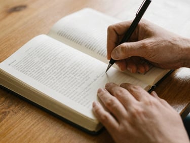 Close-up of hands writing notes in a journal next to an open Bible on a wooden table, soft morning daylight, 35mm.