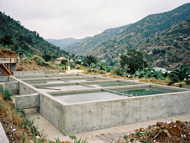 Medium-wide shot of a newly constructed concrete water filtration facility in a rural valley, natural bright overcast daylight, desaturated organic tones, medium format film, honest documentary photography.