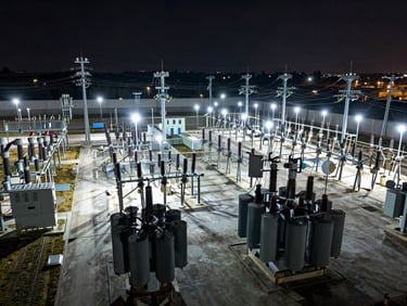 Wide overhead view of a utility substation yard at night, transformer banks and switching gear illuminated by cool white security lighting, no personnel, high-voltage equipment arranged in precise rows, dark sky above